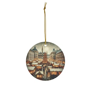 May include: Circular ceramic Christmas ornament depicting a European Christmas market in winter. The ornament shows a large Christmas tree, market stalls, and buildings. A gold string is attached for hanging.