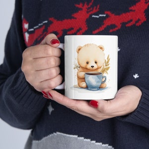 Könnte beinhalten: Weiße Keramik-Tasse mit einer Aquarell-Illustration eines Teddybären, der eine blaue Kaffeetasse hält. Der Bär ist hellbraun mit einem niedlichen Ausdruck. Die Tasse hat einen weißen Henkel und wird von einer Person gehalten, die einen marineblauen Pullover trägt.