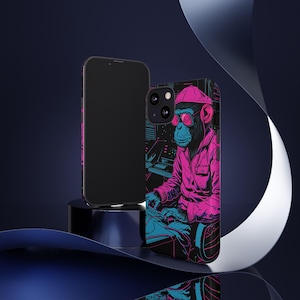May include: Black phone case with a neon illustration of a monkey in a pink hat and glasses, operating a soundboard. The design uses pink and blue colours, creating a retro pop art aesthetic. A second black phone is also visible.