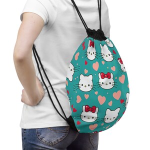 May include: A teal drawstring bag with a pattern of white cartoon cat faces with red bows and pink hearts. The bag has black drawstrings and a black base.