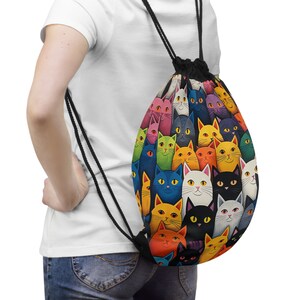 Puede incluir: Una mochila con cordón con un patrón colorido de gatos de dibujos animados en varios colores, incluyendo naranja, azul y rosa. La bolsa tiene cordones negros y una base negra. Los gatos tienen diferentes expresiones y están densamente agrupados.