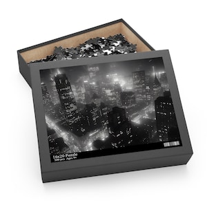 以下が含まれることがあります： モノクロのパズルボックス。都市景観の画像が描かれています。ボックスは開いており、パズルのピースが見えます。ボックスの画像は、ライトアップされた建物がある夜の都市の景色を示しています。「16x20 Puzzle」と「500 pcs Ages 9+」と記載されています。