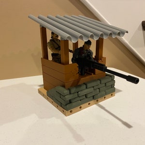 Könnte beinhalten: Ein Spielzeug-Militärposten mit zwei Figuren. Die Struktur besteht aus braunen und grauen Blöcken mit einem Wellblechdach. Eine große schwarze Waffe ragt heraus, und sandbagähnliche Blöcke bilden eine Barriere.
