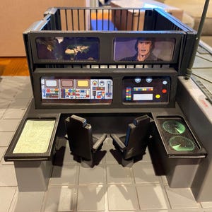 May include: A detailed view of a vintage toy command center. The gray and black structure features multiple screens displaying images and graphics, along with two black chairs. The toy includes radar screens and a map display, evoking a retro sci-fi aesthetic.