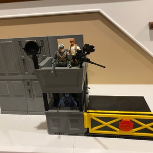Könnte beinhalten: Ein graues und schwarzes Spielzeug-Spielset mit einem grauen Turm, einer schwarzen und grauen Rampe und einem gelben Tor mit einem roten "STOP"-Schild. Der Turm hat ein kleines Fenster und einen schwarzen Geschützturm. Auf dem Turm stehen drei Spielzeugfiguren.