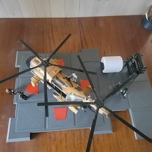 May include: A toy helicopter with camouflage paint and black blades is on a grey landing pad with red markings. The helicopter is part of a playset that includes a small fuel tank and a figure.