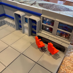 Könnte beinhalten: Ein graues und blaues Miniatur-Sci-Fi-Kommandozentrum mit zwei leuchtend orangefarbenen Stühlen. Das Set verfügt über mehrere Bildschirme mit Bildern, ein Bedienfeld und einen gefliesten Boden, ideal für Tabletop-Spiele.