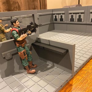 以下が含まれることがあります： 2体のフィギュアが配置されたグレーのプラスチック製アクションフィギュアプレイセット。1体のフィギュアは黒いライフルを持っています。プレイセットは、灰色のタイル張りの床と、シルエットターゲットのある灰色の壁を備えています。