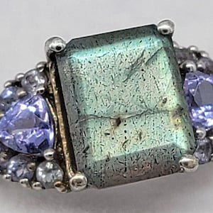 Könnte beinhalten: Ein silberner Ring mit einem großen, rechteckigen grünen Edelstein, der von kleineren, violetten Edelsteinen umgeben ist.