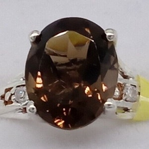 Könnte beinhalten: Ein silberner Ring mit einem großen, oval geschliffenen, dunkelbraunen Edelstein. Der Ring hat kleine, klare Akzentsteine auf beiden Seiten des Hauptsteins. Die Fassung ist ein klassisches Design.