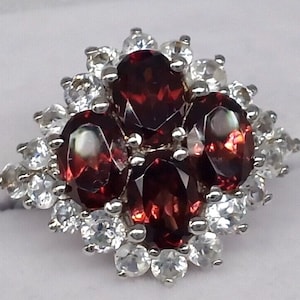 Könnte beinhalten: Ein silberner Ring mit vier ovalen, tiefroten Edelsteinen, umgeben von kleineren, runden, klaren Edelsteinen. Das Design des Rings ist ein Cluster aus Steinen, der ein Blumen- oder Sternmuster bildet. Der Ring ist vor einem weißen Hintergrund platziert.