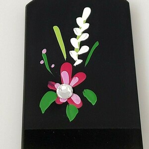 May include: Black rectangular object with a painted floral design. The design features a pink flower with a white rhinestone in the center, green leaves, and white flowers with green stems.