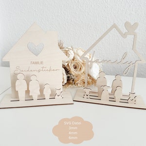 Op de afbeelding: Twee houten, huisvormige uitsnijdingen met het woord "family" en de tekst "Familie Seidenstecken" erop. De uitsnijdingen zijn versierd met harten en silhouetten van mensen en huisdieren. De uitsnijdingen bevinden zich op een wit oppervlak met een boeket gedroogde bloemen op de achtergrond.