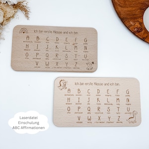 Puede incluir: Dos tableros de madera para aprender el alfabeto con el texto "Ich bin erste Klasse und ich bin" y el alfabeto. Cada tablero tiene una ilustración diferente, incluyendo un arcoíris y un dinosaurio. Los tableros son de color marrón claro.