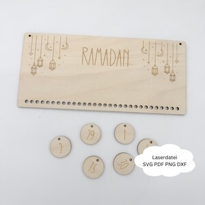 Könnte beinhalten: Ein Ramadan-Kalender aus hellem Holz mit dem eingravierten Wort "RAMADAN". Er zeigt hängende Laternen und Monde sowie kleine Holzscheiben mit den Zahlen 1, 2, 15, 29 und 30. Der Text "Laserdatei SVG PDF PNG DXF" ist ebenfalls vorhanden.