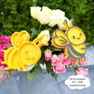 Può includere: Due decorazioni a forma di ape gialla con accenti grigi sono esposte davanti a un bouquet di rose bianche e rosa. Le decorazioni hanno un design a cartone animato. Il testo sull'immagine recita "3D Druckdatei STL + 3MF Insektentränke."