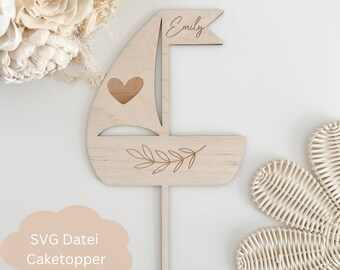 May include: A wooden sailboat cake topper with a heart and leaves engraved on it. The topper has a flag with the name "Emily" written on it.