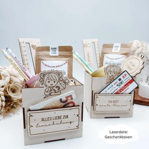Puede incluir: Dos cajas de regalo de madera llenas de útiles escolares, incluyendo lápices, reglas y pequeñas bolsas. Una caja tiene un billete de 50 euros y el texto "ALLES LIEBE ZUR Einschulung". La otra caja tiene una tarjeta de regalo y el texto "DU BIST erste Klassi".
