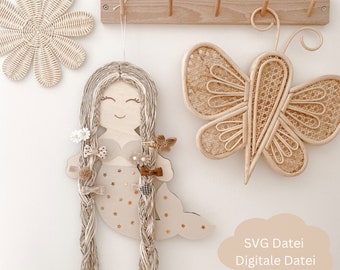 Könnte beinhalten: Eine hölzerne Meerjungfrauenfigur mit langen, geflochtenen Haaren aus Naturfaser. Die Meerjungfrau hat einen hellbraunen Körper mit einem goldenen Sternenmuster und hält eine kleine Muschel. Die Figur hängt an einer Wand mit einem Holzschmetterling und einer blütenförmigen Wanddekoration.