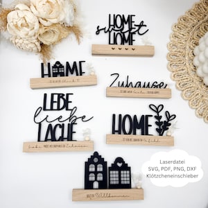Könnte beinhalten: Eine Auswahl an schwarzen Holzschildern mit Schriftzügen wie "Home Sweet Home" und "Zuhause". Jedes Schild ist auf einem hellen Holzsockel montiert. Die Schilder sind mit kleinen floralen Akzenten verziert.