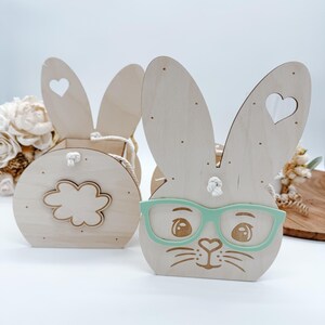 May include: Two wooden bunny shaped Easter baskets. One basket is a simple bunny shape with a heart cut out of each ear. The other basket has a bunny face with green glasses and a cloud shaped cut out on the back.