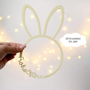 Op de afbeelding: Een lichtbeige paashaas oordecoratie met de woorden "Frohe Ostern" in een cursief lettertype. De decoratie wordt gehouden voor een achtergrond van zachte, onscherpe lichten. Een witte, wolk-vormige tekstballon leest "3D Druckdatei STL 3MF".