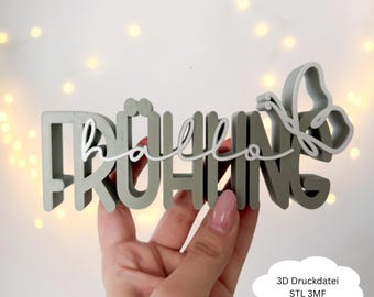 May include: A gray 3D-printed sign that reads "Frühling" with "hello" in white script. A butterfly design is attached to the end of the word. The sign is held in a hand.
