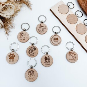 Könnte beinhalten: Eine Sammlung von Holzschlüsselanhängern mit silbernen Schlüsselringen. Die Schlüsselanhänger weisen eingravierte Designs auf, darunter Pfotenabdrücke, Menschen mit Haustieren und Texte wie "Dog Mom" und "Best Grandma". Die Schlüsselanhänger sind rund und aus hellem Holz gefertigt.