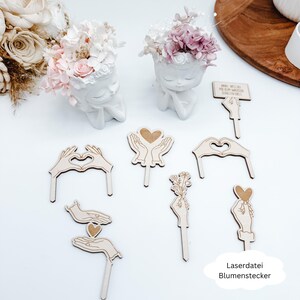 May include: A collection of wooden flower stakes with heart and hand designs, alongside two white ceramic head planters filled with pink and white flowers. The stakes feature various hand gestures and floral arrangements. The text "Laserdatei Blumenstecker" is visible.