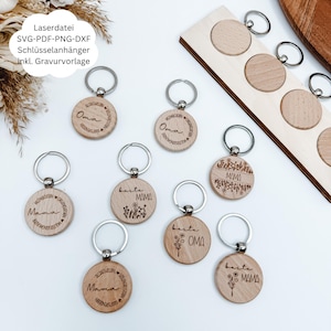 Könnte beinhalten: Holz-Schlüsselanhänger mit silbernen Schlüsselringen. Die Schlüsselanhänger sind rund und mit den Wörtern "Oma" und "Mama" sowie floralen Designs graviert. Ein Holzhalter mit kreisförmigen Aussparungen ist ebenfalls sichtbar. Der Text "Laserdatei SVG-PDF-PNG-DXF Schlüsselanhänger inkl. Gravurvorlage" befindet sich im Bild.