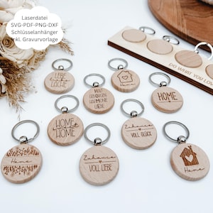 Puede incluir: Una colección de llaveros de madera con anillas plateadas. Los llaveros presentan diseños y textos grabados, incluyendo patrones florales, casas y frases como "Home" y "Love". Los llaveros son redondos y están hechos de madera clara.
