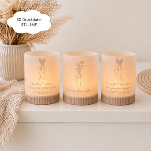May include: Three illuminated frosted glass candle holders with beige bases. Each features a different design: a hand holding a heart, a hand holding flowers, and a hand holding a heart. Text in German is printed on each holder.