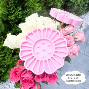 Puede incluir: Se muestran dos objetos rosas con forma de flor, impresos en 3D, uno más grande que el otro. El más grande está en primer plano y el más pequeño en segundo plano. Están colocados sobre un fondo de rosas rosas y blancas y vegetación. El texto de la imagen dice "3D Druckdatei STL + 3MF Insektentränke."