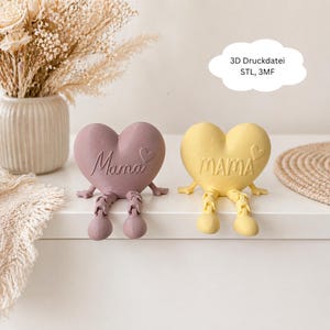 Pode incluir: Duas figuras em forma de coração, uma malva e outra amarela, cada uma com pernas articuladas. A palavra "Mama" está inscrita em cada coração. Uma bolha de texto em forma de nuvem diz "3D Druckdatei STL, 3MF".