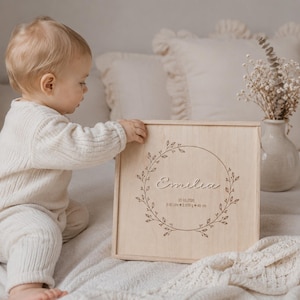 Könnte beinhalten: Eine quadratische Erinnerungsbox aus hellem Holz mit einer lasergravierten, kreisförmigen Girlande. In der Mitte steht der Name Emilia in Schreibschrift, gefolgt vom Datum 20.03.2026, der Uhrzeit 0:30 Uhr, dem Gewicht 2.830 g und der Größe 48 cm.