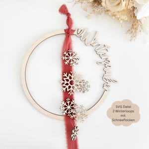 Puede incluir: Un aro de madera con la palabra "Winterzauber" escrita en él. Una cinta roja cuelga del aro con copos de nieve de madera unidos a ella. El texto "SVG Datei 2 Winterloops mit Schneeflocken" está escrito sobre un fondo blanco.