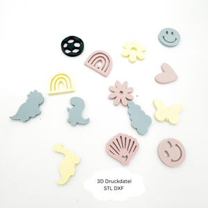 May include: A collection of pastel-colored 3D-printed shapes, including dinosaurs, rainbows, flowers, a butterfly, a seashell, and a smiley face. The shapes are scattered on a white surface. Text at the bottom reads "3D Druckdatei STL DXF."