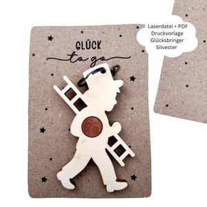 Puede incluir: Un amuleto de madera de deshollinador con una escalera y una moneda, sujeto a una tarjeta marrón con las palabras "Glück to go" e ilustraciones de estrellas. El amuleto es de color beige claro.