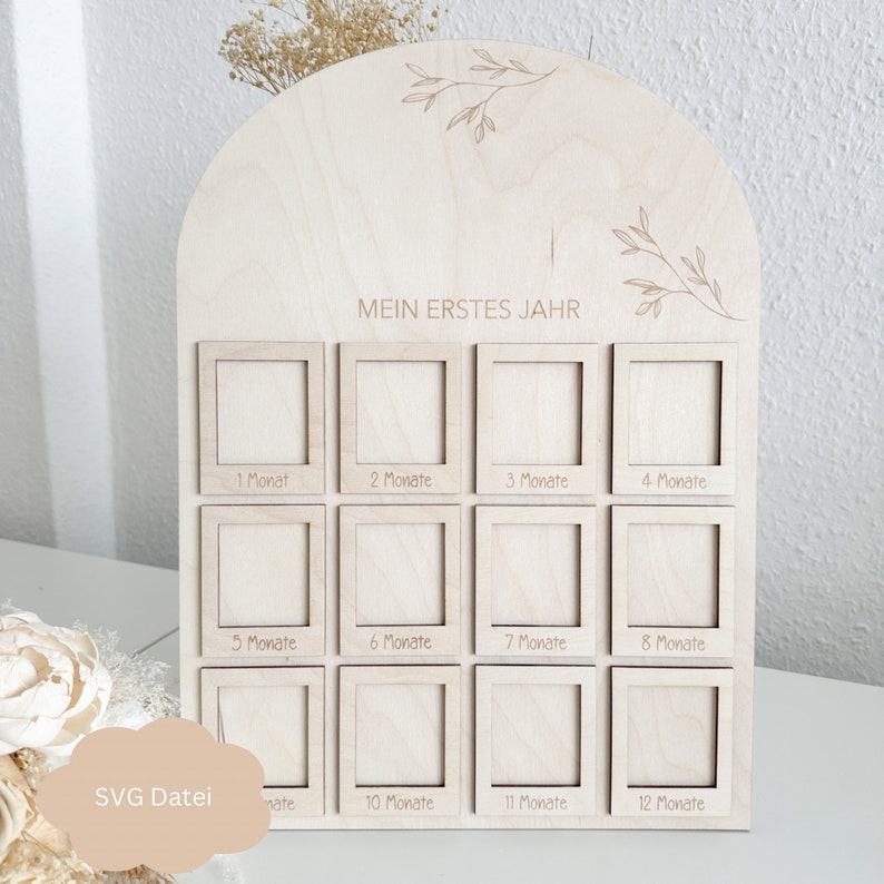 May include: A wooden photo frame with 12 squares for pictures, each labelled with a month of the year in German. The frame is decorated with a floral design and the text "Mein erstes Jahr" (My first year).