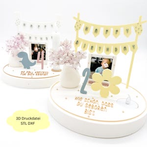 May include: White cake toppers with a photo frame, flowers, and decorative elements. One has a blue dinosaur and the number 4, the other has a yellow flower and the number 2. Both have a banner that says "Happy Birthday".