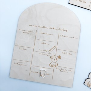 Puede incluir: Una placa de madera con una mancha marr&oacute;n claro, que presenta una tabla para registrar los hitos del primer cumplea&ntilde;os de un ni&ntilde;o. La tabla incluye secciones para las primeras palabras del ni&ntilde;o, su peso, su altura y otros logros. La placa est&aacute; decorada con un peque&ntilde;o conejito de madera y un diente.