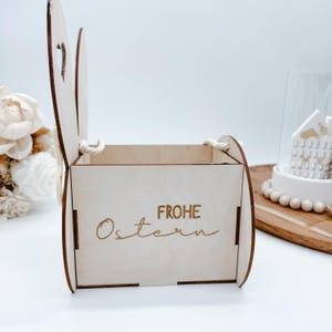 May include: A wooden Easter bunny basket with a white rope handle. The basket is decorated with the words "Frohe Ostern" in gold lettering.