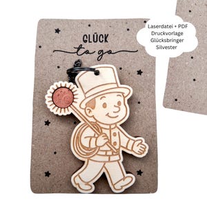 May include: A wooden chimney sweep keyring with a coin, a top hat, and a coil of rope. The keyring is attached to a brown card with the words "Glück to go" and star shapes. The keyring is a good luck charm.