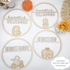 Könnte beinhalten: Vier runde Holzschilder mit lasergeschnittenem Text und Illustrationen. Die Schilder zeigen Sätze wie "Herbstlich Willkommen" und "Herbstzauber". Jedes Schild hat eine andere herbstliche Illustration, wie einen Igel oder einen Bären. Die Schilder sind aus hellem Holz gefertigt.