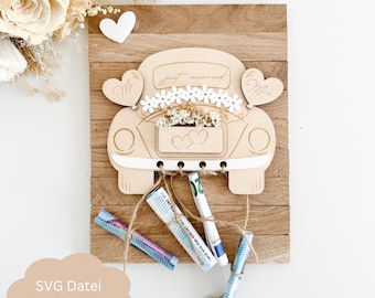 May include: Wooden wedding card holder shaped like a car with the words "just married" on the back window. The car has white hearts on the sides and a floral garland on the top. The car is attached to a wooden background with twine and money is tied to the twine.