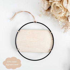 Pode incluir: Uma moldura redonda de metal preto com uma placa de madeira de cor clara. Um cordão de couro bege é preso para pendurar. O texto "SVG Datei digitale Datei" está em uma nuvem marrom claro no canto inferior esquerdo. Flores secas ao fundo.