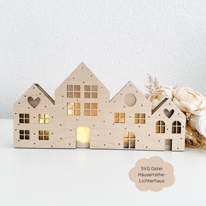 May include: A wooden light-up house design with four houses, each with windows and a heart cutout. The houses are connected and have a dotted pattern. The text "SVG Datei Häuserreihe-Lichterhaus" is in the bottom right corner.