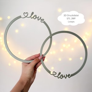 Könnte beinhalten: Zwei graue kreisförmige Wanddekorationen mit dem Wort "love" und einem Herzdesign. Der Text "3D Druckdatei STL 3MF Loops" befindet sich in einer Wolke. Die Dekorationen werden von einer Hand gehalten.