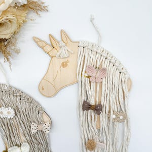 Pu&ograve; includere: Testa di unicorno in legno con criniera e coda di filo bianco. L'unicorno ha una farfalla rosa, un fiocco marrone e mollette per capelli a forma di fiore bianco attaccate alla criniera.