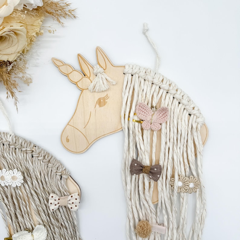 Pu&ograve; includere: Testa di unicorno in legno con criniera e coda di filo bianco. L'unicorno ha una farfalla rosa, un fiocco marrone e mollette per capelli a forma di fiore bianco attaccate alla criniera.